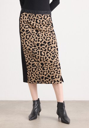 Femme portant une jupe midi à imprimé léopard avec des panneaux latéraux noirs et des bottines noires, debout sur un sol gris.