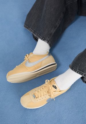 Baskets Nike beiges avec un swoosh argenté, portées avec des chaussettes blanches et un jean noir, pieds reposant sur un sol moquetté bleu.