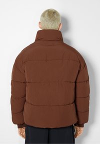 Giacca a vento marrone con colletto alto e trapuntatura orizzontale. Presenta lacci regolabili all'orlo e una texture di tessuto liscia.