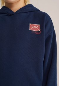 Tums zilas kapuce, kas izgatavota no mīkstas auduma, ar sarkanu un baltu aploksnes grafiku, kurā redzamas sirds ikona un pasta zīmogs detaļas uz krūtīm.