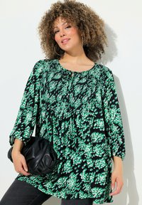 Tunic nera con motivo floreale verde, colletto rotondo, dettagli arricciati, maniche a tre quarti, abbinata a una borsa nera. Tessuto liscio.