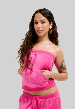 Jonge vrouw met lang krullend haar, gekleed in een roze strapless top en bijpassende broek, toont een vlindertattoo op haar bovenarm.