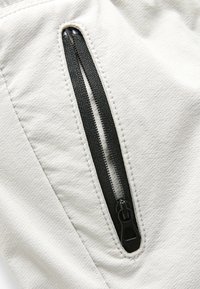 Cerniera verticale nera parzialmente aperta su tessuto grigio chiaro testurizzato con cuciture visibili intorno all'apertura della cerniera.