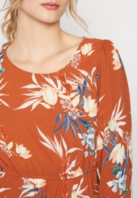 Hüftfarbendes Kleid mit floralem Muster, das einen runden Halsauschnitt und lange Ärmel hat. Der Stoff hat eine strukturierte Oberfläche mit gerafften Akzenten.