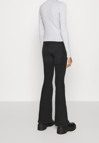 Henkilö, jolla on päällään mustat levenevät farkut, mustat möhkäleplatfformikengät ja vaaleanharmaa pitkähihaisen ribbipaita, seisoo valkoista taustaa vasten.