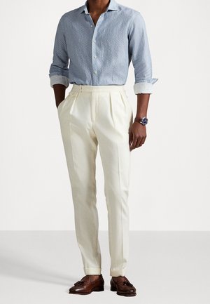 Man wearing light blue textured button-up shirt with rolled sleeves, high-waisted cream pleated trousers, brown leather tassel loafers, and watch.
