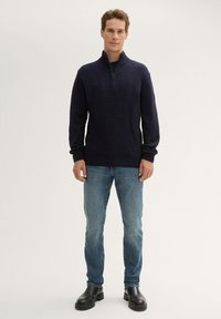 Marineblauwe gebreide trui met een ritskraag, geribbelde manchetten en een losse pasvorm, gecombineerd met lichtblauwe jeans en zwarte laarzen.
