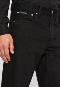 Jeans de mezclilla negra con un ajuste ajustado, que cuentan con un bolsillo frontal, herrajes plateados y una etiqueta de Calvin Klein en la cinturilla.