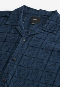Camisa azul marino abotonada con un patrón cuadrado texturizado y bordado y un cuello. Cuenta con cuatro botones y una etiqueta de marca en el interior.