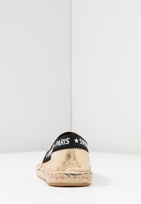 Espadrille con parte superior de ante dorado, banda elástica negra con texto blanco "SUPERGA PARIS"; entresuela envuelta en yute y suela de goma.