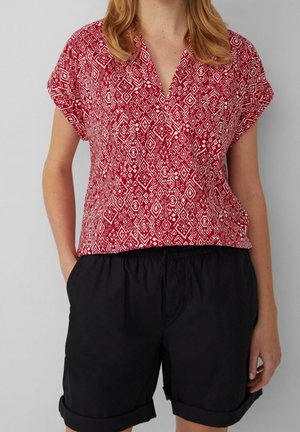 Femme portant un chemisier rouge à manches courtes à motifs et un short noir retroussé, debout les mains dans les poches.