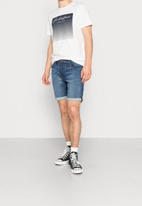 Muž v bílém tričku s grafikou, vyhrnutých modrých denimových šortkách, bílých ponožkách a černých teniskách s vysokou špicí stojí před jednobarevným pozadím.