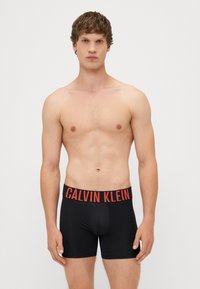 Boxeri negri cu talie groasă de culoare roșie, având inscripția "CALVIN KLEIN" în litere îngroșate. Textură netedă și design ajustat.