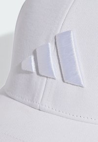 Biely klobúk vyrobený z textúrovanej tkaniny má tri biele vyšívané akcenty v geometrickom tvare na prednej strane. Šitie je viditeľné pozdĺž okraja.