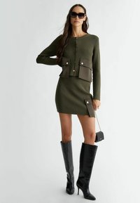 Cardigan a costine verde oliva con due tasche frontali, abbinato a una mini gonna coordinata. Stivali neri lucidi fino al ginocchio e una piccola borsa completano il look.