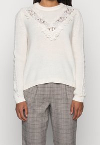 Pull blanc en tricot avec un motif en dentelle florale au niveau de l'encolure, tissu texturé et poignets côtelés. Porté avec un pantalon à carreaux.