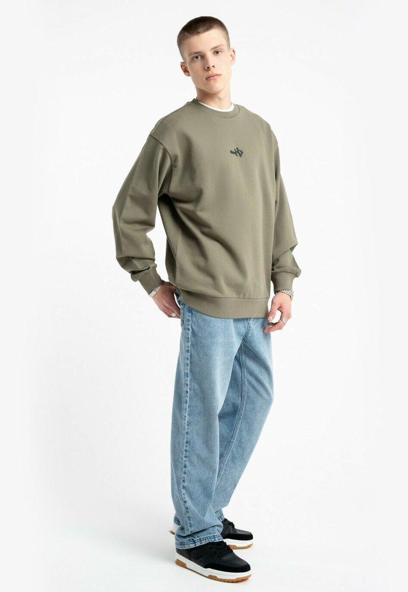 Młody mężczyzna stoi z rękoma w okolicy kieszeni, ubrany w oliwkowy sweter, jasnoniebieskie szerokie dżinsy, czarne sneakersy i srebrne akcesoria na nadgarstku.