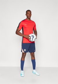 Camiseta de fútbol roja de manga corta con detalles en negro, pantalones cortos azul marino con el logo del equipo, calcetas azules y zapatillas de fútbol azul claro, sosteniendo un balón de fútbol.
