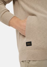 Beige sweater with a knit texture and ribbed cuffs. Features a side pocket and a black label stating "MADE FOR DAILY ADVENTURES."