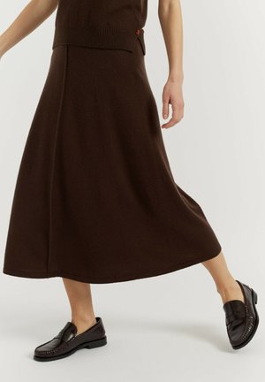 Vrouw die een bruine midi-rok en zwarte leren loafers draagt, lopend met één been naar voren op een lichte achtergrond.