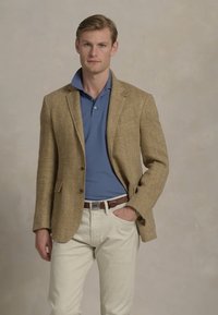 Blazer castanho em padrão de espinha de peixe com lapelas de bico, usado sobre uma camisa polo azul e calças brancas, complementado por um cinto de couro castanho.