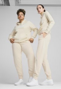 Beige hoodies en joggingsbroeken van zachte stof, voorzien van een kangaroo pocket en logodetail. Witte sneakers maken de outfit compleet.