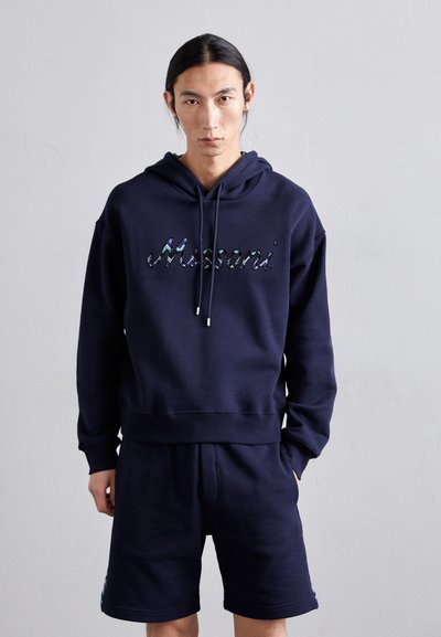Haut à capuche bleu marine avec un cordon de serrage, arborant un logo texturé et irisé. Assorti avec un short bleu marine, également de la même teinte. Coupe décontractée.