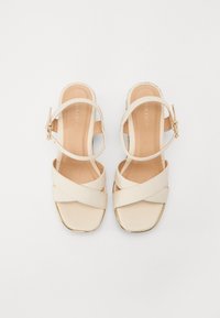 Beige cross-strap sandals with a square toe and a gold-accented block heel. Adjustable ankle strap with a metallic buckle. Smooth texture.