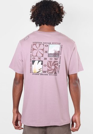 Homme aux cheveux bouclés portant un T-shirt mauve avec un graphisme en quatre parties et le texte « "European Surfing Company Oxbow" » au dos.