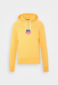 Žlutá mikina s kapucí, přední kapsou na břiše, nastavitelným stahovacím šňůrem a vyšívaným štítovým logem v červené, bílé a modré barvě.