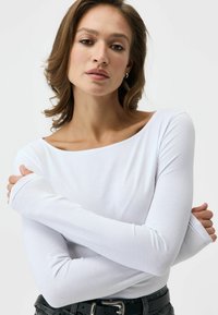 Długi rękaw, kremowa bluzka wykonana z miękkiego materiału, z szerokim dekoltem i dopasowanymi rękawami. Zestawiona z czarnym paskiem i teksturowanym dżinsem.