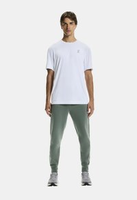 Witte t-shirt met korte mouwen met een klein logo, gecombineerd met groene joggingsbroeken en grijze sportschoenen, staande tegen een witte achtergrond.