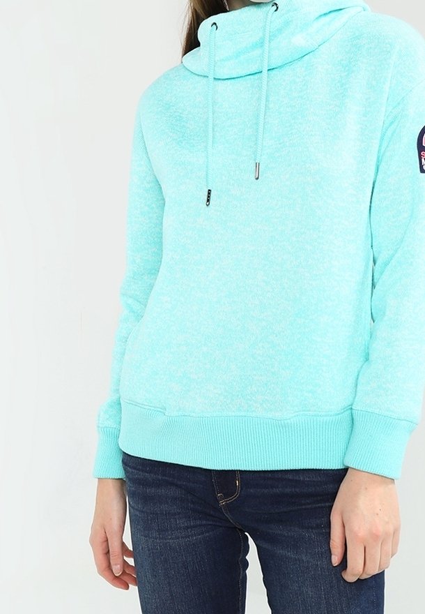 Personne portant un sweat à capuche bleu clair avec cordons et un jean bleu foncé, debout devant un fond blanc uni.