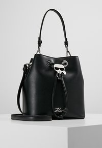 Sort læder bucket-taske med snøre lukning, der har en sølv logo og en dekorativ vedhæng med en figur iført solbriller.