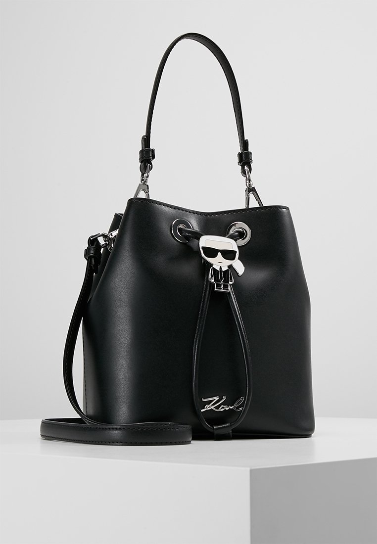 Sort læder bucket-taske med snøre lukning, der har en sølv logo og en dekorativ vedhæng med en figur iført solbriller.