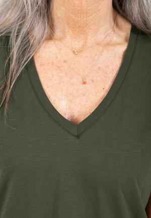 Olivgrünes V-Ausschnitt-T-Shirt mit weicher Textur. Zwei zarte goldene Halsketten in Lagen, eine mit einem Herz und eine mit einer kleinen orangenen Perle.