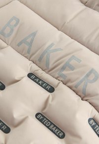 Gepolsterte Jacke aus hellem beigem Stoff mit erhabenem "BAKER"-Text in Silber und Marken-Patches in dunkler Farbe mit glänzenden Akzenten.