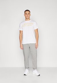 Witte T-shirt met gele grafische tekst, gecombineerd met grijze sweatpants en witte sneakers. Eenvoudig ontwerp, casual pasvorm, zachte katoenen textuur.