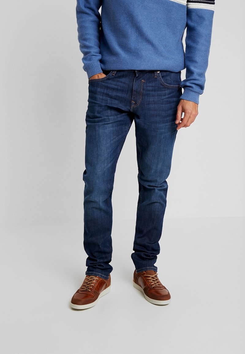 Donkerblauwe denim jeans met een rechte pasvorm, voorzien van subtiele vervaging, vijf zakken en gestikte details. Gecombineerd met bruine sneakers.