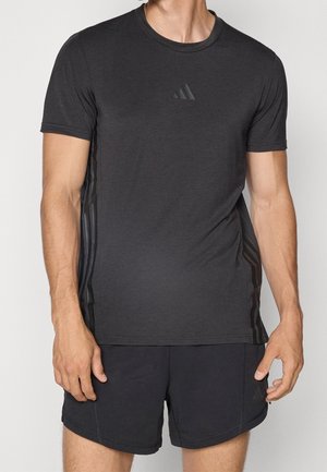 Man draagt een zwarte atletische T-shirt en korte broek met subtiele Adidas-logo's en strepen aan de zijkanten, staand tegen een effen achtergrond.
