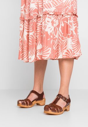 Brune lædertræsko med åben tå og rem, der har en træbund og dekorativ syning. Sættes sammen med en lyserød blomsterkjole.