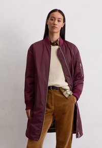 Bordeaux lange bomberjack met ribgebreide manchetten, ritsdetails, gelaagd over een crèmekleurige trui en een geruite overhemd, gecombineerd met een bruine corduroy broek.