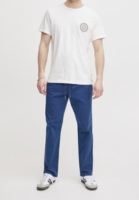 Biała bawełniana koszulka z małym okrągłym logo na piersi, w zestawie z niebieskimi bawełnianymi spodniami i białymi tenisówkami z szarymi akcentami.