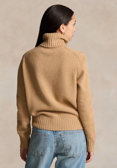 Hosszú sötét hajú nő, aki teve színű bordázott garbót és világoskék farmert visel, háttal egy egyszínű háttér előtt.