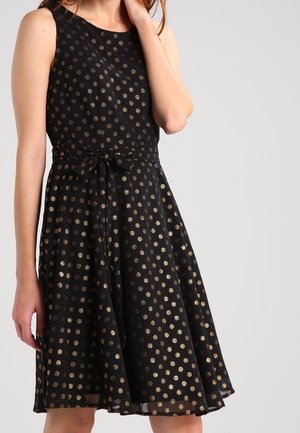 Ærmeløs sort kjole med metallic guldprikker og bælte bundet i sløjfe ved taljen, båret af en stående kvinde.