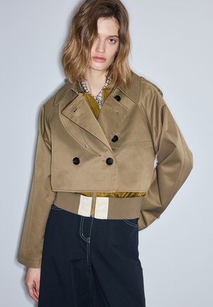 Vrouw met halflang lichtbruin haar draagt een gelaagde, cropped beige jas, geruit overhemd en donkere broek, staand voor een effen grijze muur.