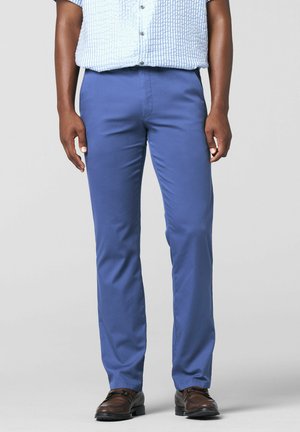 Man draagt blauwe rechte broek, een wit gestructureerd overhemd met korte mouwen en bruine leren nette schoenen, staand tegen een effen achtergrond.