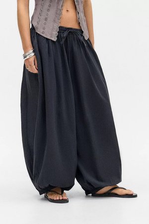 Personne portant un pantalon harem noir ample avec des ourlets élastiques, un haut gris sans manches, des bracelets argentés et des sandales noires en tongs, debout sur un sol blanc.