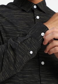 Camicia nera con delicato motivo a strisce dorate. Colletto con bottoni, bottoni bianchi sui polsini e tessuto con finitura testurizzata.