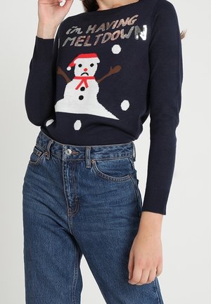 Granatowy sweter z grafiką topniejącego bałwana i napisem „I'm having meltdown”, noszony z wysokim stanem niebieskich dżinsów, swobodna poza.